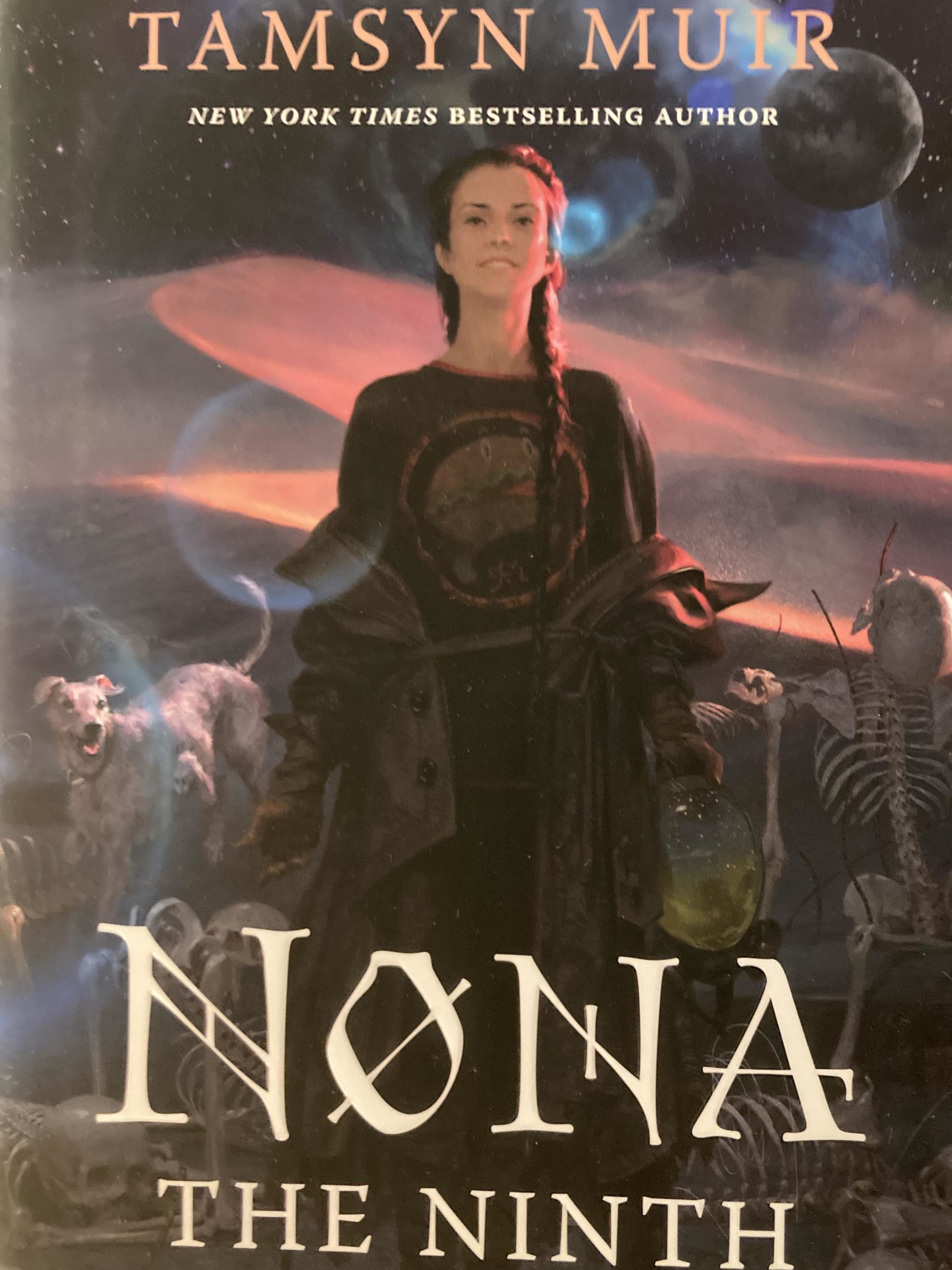 The cover of NONA THE NINTH. A dark-haired woman in braids with a hamburger on her t-shirt stands, smiling, in a field of skeletons in tortured poses. A white dog with six legs is in the background.