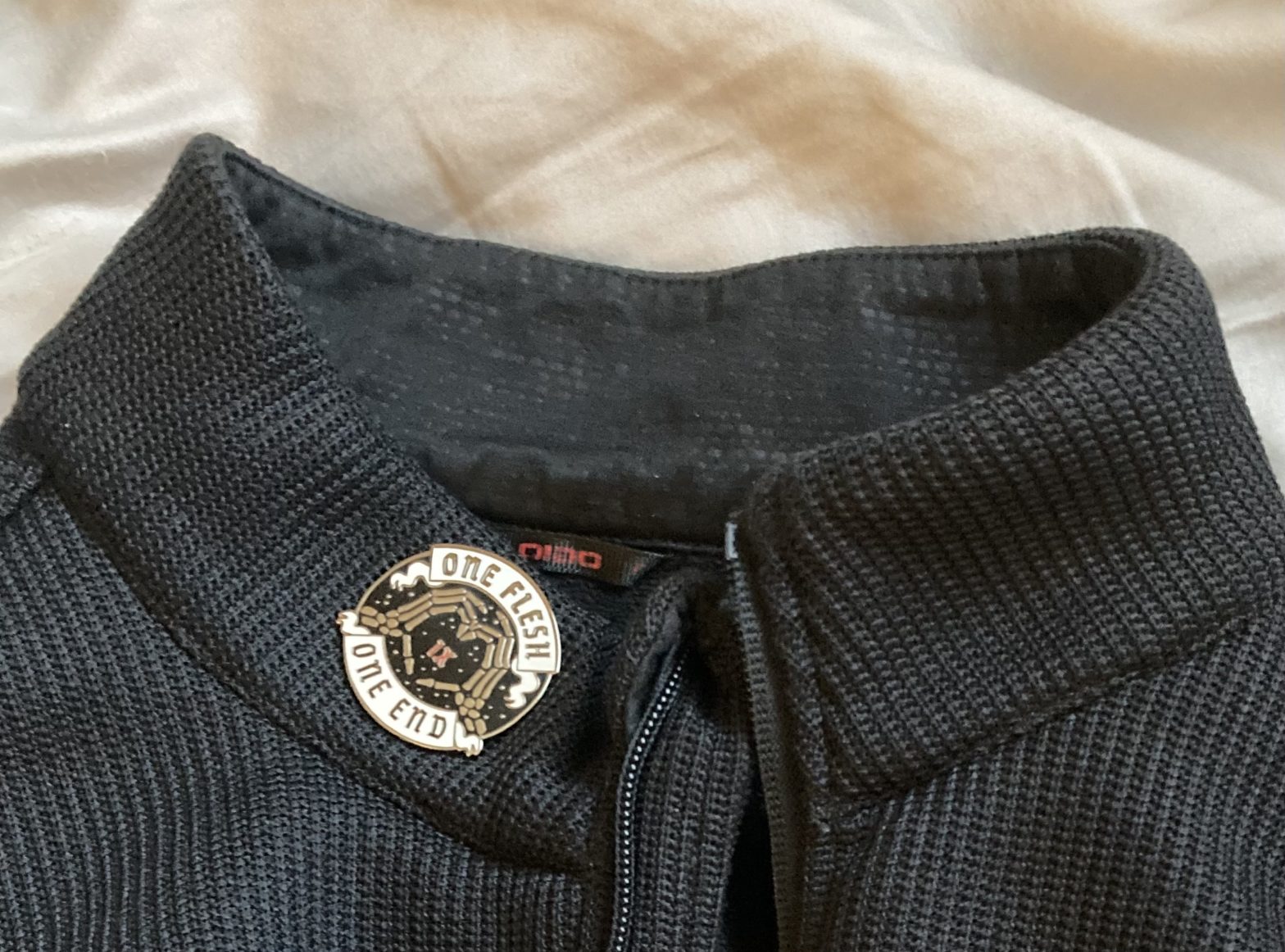 An enamel pin where two skeletal hands make a heart shape around the Roman numeral IX. A scroll at the top says "One flesh, one end."